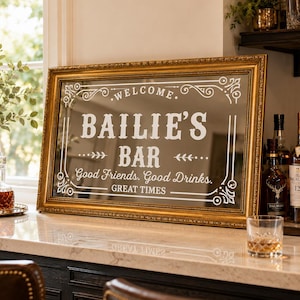 Pode incluir: Um espelho retangular com moldura dourada apresentando texto branco que diz WELCOME BAILIE'S BAR Good Friends, Good Drinks, GREAT TIMES. O espelho é decorado com bordas de filigrana branca e está posicionado sobre uma bancada branca.