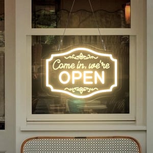 Op de afbeelding: Een wit bord met zwarte letters met de tekst "Come in, we're open" hangt in een winkelruit. Het bord is verlicht met witte LED-verlichting. Twee kleine tafels en stoelen staan voor de winkel.