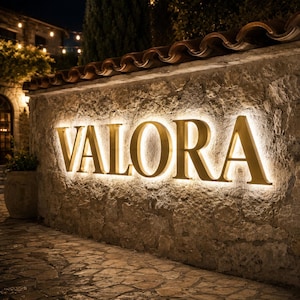 May include: A large, gold-coloured, three-dimensional sign spelling the word VALORA mounted on a textured, light-coloured stone wall. The letters are backlit, creating a glowing halo effect against the rough surface of the masonry wall.