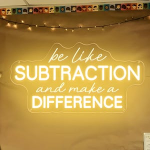 May include: A classroom scene with a brown bulletin board featuring a neon sign that reads "be like SUBTRACTION and make a DIFFERENCE." A wooden bench with a striped pillow sits below the sign. An alphabet banner hangs to the left.