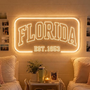 Może przedstawiać: Prostokątny neon zamontowany na ścianie z napisem FLORIDA dużymi, pogrubionymi literami z białym obrysem oraz tekstem EST. 1853 poniżej. Neon emituje ciepłe, jasne światło, które oświetla przestrzeń sypialni.