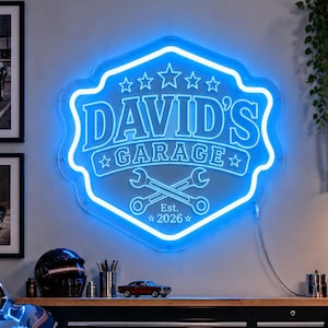 May include: A bright blue neon sign shaped like a shield with the text DAVID'S GARAGE, five stars, and two crossed wrenches. Below the wrenches, the text reads Est. 2026. The sign is mounted on a wall above a black metal tool cabinet.
