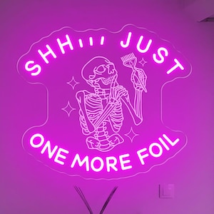 May include: A pink neon sign in a salon. The sign displays a skeleton holding a brush, with the words "SHHH JUST ONE MORE FOIL". The sign is mounted on a white wall, with a small table and chair visible.
