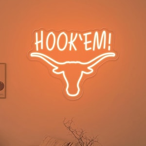 May include: Orange neon sign with a white outline of a longhorn steer head and the words "HOOK'EM!" above the head. The sign is hanging on a wall in a living room with a couch, coffee table, and artwork.