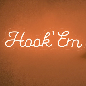 May include: A neon sign that reads "Hook 'Em" in a cursive font, mounted on a wall. A brown football sits on a small shelf, and a framed picture hangs nearby. A beige sofa with a brown pillow is in the foreground.