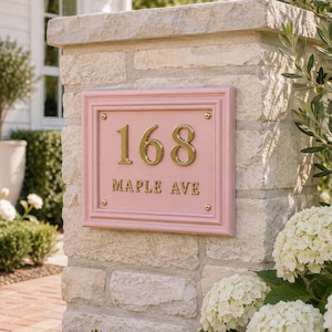 Pode incluir: Uma placa retangular rosa de número de casa com uma moldura em relevo, apresentando os números 168 em dourado e o texto MAPLE AVE em letras douradas. A placa é montada com quatro parafusos dourados em um pilar de pedra de cor clara.