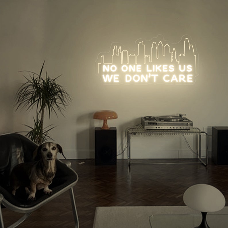 May include: A white neon sign with a cityscape silhouette and the text "NO ONE LIKES US WE DON'T CARE" on a white wall. A small dog sits on a black chair in the foreground.