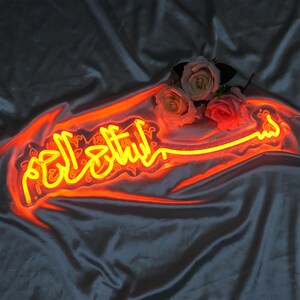May include: A neon sign with the Arabic phrase "Subhanallah" in yellow neon lettering on a red background. The sign is illuminated and is sitting on a dark gray fabric surface.