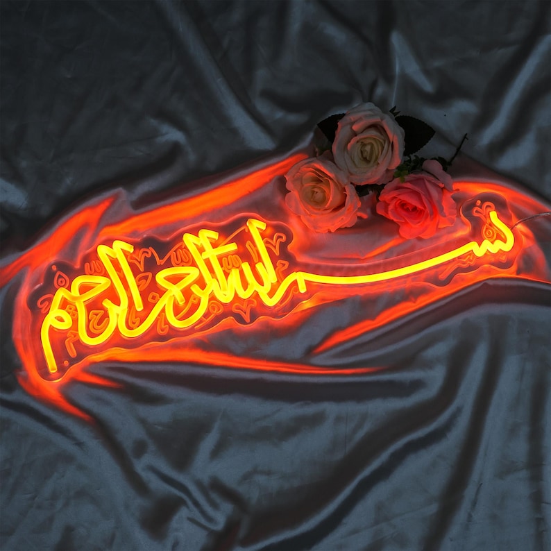 May include: A neon sign with the Arabic phrase "Subhanallah" in yellow neon lettering on a red background. The sign is illuminated and is sitting on a dark gray fabric surface.