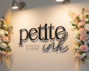 May include: A wall-mounted sign that reads "petite ink STUDIO & ACADEMY" in black lettering. The sign is centered on a light-colored wall, flanked by floral arrangements with pink and white flowers. Two silver wall sconces provide illumination.