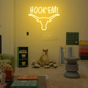 May include: A yellow neon sign with the words "HOOK'EM!" and a stylized longhorn steer head. The sign is hanging on a wall in a room with a dog sitting on a checkered cushion.