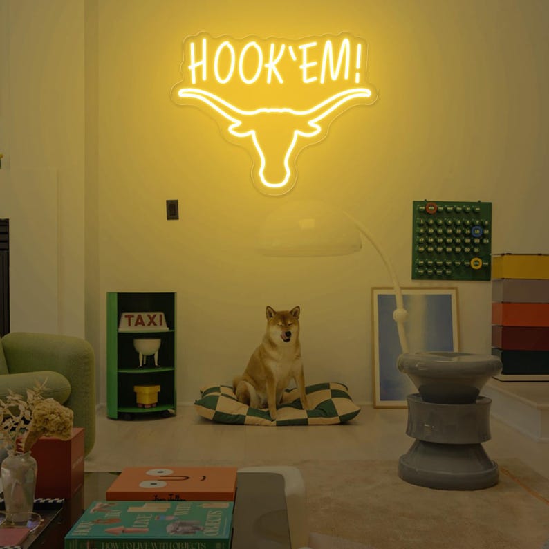 May include: A yellow neon sign with the words "HOOK'EM!" and a stylized longhorn steer head. The sign is hanging on a wall in a room with a dog sitting on a checkered cushion.