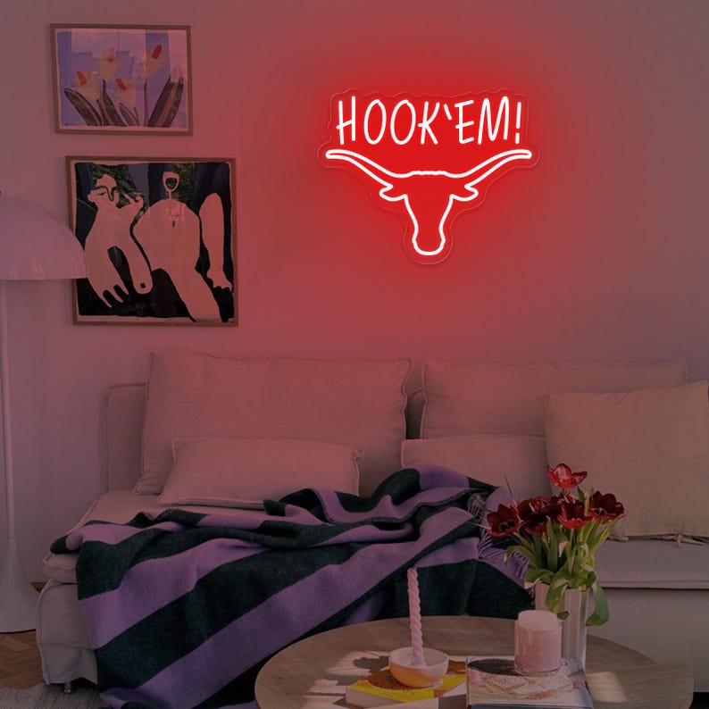 May include: Red neon sign with a white outline of a longhorn steer head and the text "HOOK'EM!"  The sign is mounted on a white wall above a couch with a purple and gray striped throw blanket.