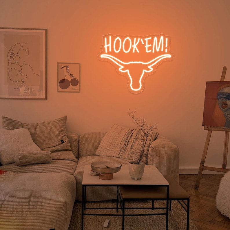 May include: Orange neon sign with a white outline of a longhorn steer head and the words "HOOK'EM!" above the head. The sign is hanging on a wall in a living room with a couch, coffee table, and artwork.