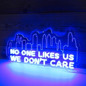 May include: White neon sign with a city skyline silhouette and the text "NO ONE LIKES US WE DON'T CARE". The sign is lit up in a bright blue colour.