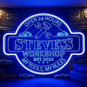 Può includere: Un'insegna al neon blu con la scritta "STEVES WORKSHOP" e "OPEN 24 HOURS". L'insegna presenta una chiave inglese e un martello, insieme al testo "MY TOOLS, MY RULES" e "EST.2023". Sono visibili modellini di auto.