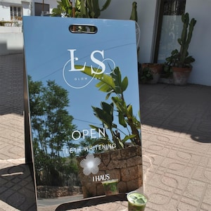 Pode incluir: Uma placa publicitária espelhada com o texto "LS GLOW BAR" e "OPEN TEETH WHITENING". A placa reflete o céu, as árvores e um edifício. Uma pequena floreira de pedra com vegetação está presa à placa.