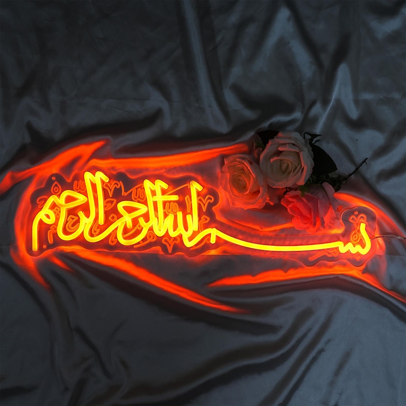 May include: A neon sign with the Arabic phrase "Bismillah" in yellow lettering on a white background. The sign is illuminated with a warm orange glow.