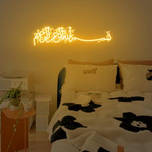 May include: A bedroom with a bed with a white and black patterned blanket. The bed has a white headboard and two white pillows with a brown dog design. There is a neon sign on the wall that says "Allah" in Arabic.