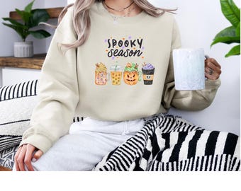 May include: Beige sweatshirt with the text "Spooky Season" above a row of four Halloween-themed drinks. The drinks include a pumpkin spice latte, a caramel apple cider, a pumpkin-shaped drink, and a purple drink.