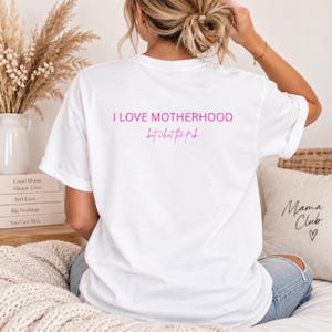 Könnte beinhalten: Ein weißes kurzärmeliges T-Shirt mit rosafarbenem Text auf der Rückseite, der I LOVE MOTHERHOOD in einer fetten serifenlosen Schrift zeigt, gefolgt von dem Satz but what the f*ck in einer kleineren Schreibschrift, mittig auf dem oberen Rücken.