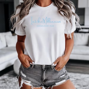 Könnte beinhalten: Ein weißes kurzärmeliges T-Shirt mit dem hellblauen Schriftzug Into the Millennium und DÜSSELDORF 2026 auf der Brust, kombiniert mit einer grauen, abgenutzten Jeansshorts mit ausgefransten Säumen und sichtbarem weißem Taschenfutter.