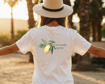 May include: White t-shirt with a watercolor lemon and leaf design, and the words "Limoncello Spritz Club" in a script font. A person is wearing a beige hat with a black band. The background is blurred with sunlight.