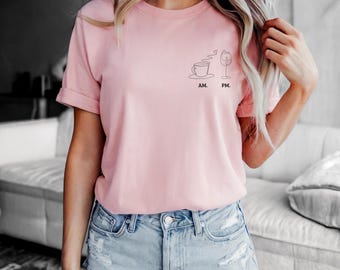 May include: A light pink short-sleeve t-shirt featuring a black line art graphic on the left chest. The design depicts a coffee cup labeled AM and a wine glass labeled PM, with steam rising from the cup. The shirt has a crew neckline and rolled sleeves.