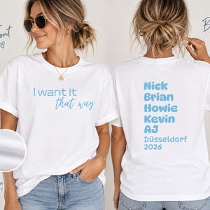 Könnte beinhalten: Ein weißes kurzärmeliges T-Shirt mit dem hellblauen Text I want it that way auf der Vorderseite. Die Rückseite des Shirts zeigt die hellblauen Namen Nick, Brian, Howie, Kevin und AJ, gefolgt von dem Text Düsseldorf 2026 in Hellblau.