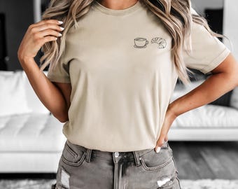 May include: A beige short-sleeve t-shirt featuring a small black line art graphic on the left chest area. The graphic depicts a simple coffee cup on a saucer positioned next to a croissant. The shirt is paired with distressed grey denim shorts.