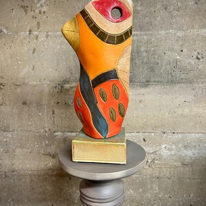 May include: Abstract ceramic sculpture in orange, red, blue, and brown with a hole in the center of the eye. The sculpture is on a square base and a gray pedestal.
