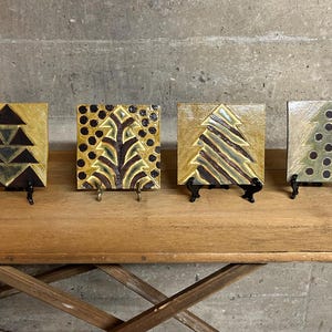 May include: Four ceramic tiles with gold and black geometric designs of stylized Christmas trees. The tiles are displayed on small black stands on a wooden surface.