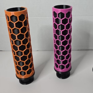 May include: Three cylindrical tubes with a black inner core and an outer honeycomb lattice shell. The outer shells are orange, pink, and black. Each tube features a circular base attachment at the bottom, standing upright in a row.