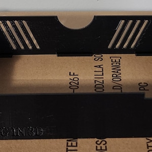 May include: Black plastic 3D printed box with a rectangular opening and a small cutout on the top. The box has a label on the bottom that reads "JRHETING IN 3D" and a label on the side that reads "ODZILLA SO -026 F LD/ORANGE] PC".