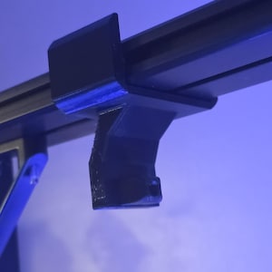 May include: Close-up of a black plastic bracket attached to a black metal rail. The bracket has a curved design and appears to be 3D-printed. The background is a gradient of blue.