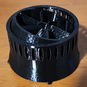 May include: Black plastic 3D printed object with a circular base and a central fan-like structure with multiple blades. The object has a series of small holes around the base.