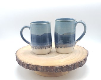 May include: Two handmade ceramic mugs with a blue and white gradient design. The mugs have a curved handle and are displayed on a wooden cake stand. The mugs are perfect for coffee, tea, or any hot beverage.