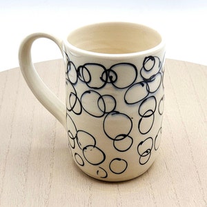 May include: A white ceramic mug with a black and white abstract design of circles. The mug has a handle.