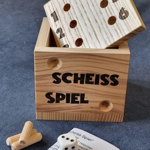 Könnte beinhalten: Holzwürfelspiel mit einer Box und einem Deckel. Die Box ist mit "SCHEISS SPIEL" in schwarzen Buchstaben beschriftet. Der Deckel hat Zahlen und Punkte. Enthält einen weißen Würfel, Holzstifte und ein gedrucktes Anleitungsblatt.