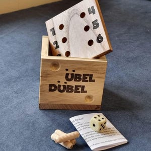 Könnte beinhalten: Holzspiel Übel Dübel. Das Set enthält eine Holzkiste mit dem Namen des Spiels, einen Holzspielwürfel mit Zahlen und einen Holzblock mit Löchern. Ein kleiner weißer Würfel und Anweisungen sind ebenfalls enthalten.