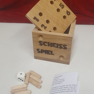 Könnte beinhalten: Ein Holzspielkasten mit der Aufschrift "SCHEISS SPIEL" auf der Vorderseite. Die Box hat einen Deckel mit Löchern, die von 1 bis 6 nummeriert sind. Das Spiel beinhaltet einen weißen Würfel mit schwarzen Punkten und Holzstäbchen. Die Anleitung ist auf einer weißen Karte gedruckt.