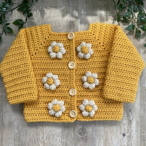 May include: A mustard yellow crocheted cardigan with white daisy appliques and wooden buttons.