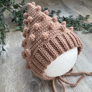 May include: A handmade, light brown crochet bonnet with a textured, bobble stitch design. The bonnet has a ribbed brim and ties for securing. The bonnet is displayed on a white, spherical form.