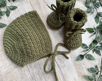 May include: A set consisting of a crocheted olive green bonnet with long ties and a pair of matching crocheted olive green booties. The items feature a textured yarn construction with a ribbed cuff design on the booties and a rounded shape.