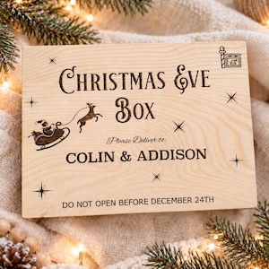 Könnte beinhalten: Eine rechteckige, helle Holzbox mit einer Lasergravur, die den Text CHRISTMAS EVE BOX, Please Deliver to: COLIN & ADDISON und DO NOT OPEN BEFORE DECEMBER 24TH sowie Illustrationen eines Weihnachtsmanns im Schlitten und Sternen zeigt.