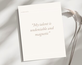 May include: A cream-colored affirmation card with the text "My talent is undeniable and magnetic" in a soft, neutral font. The word "Affirmation" is printed at the top. The card is placed on a white surface with a dried floral arrangement.