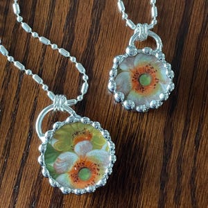 Può includere: Due collane d'argento con ciondoli floreali. I ciondoli sono rotondi e presentano un fiore bianco con accenti arancioni e verdi.