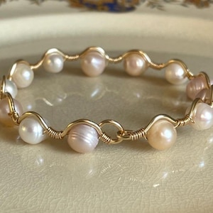 May include: A gold-toned bracelet featuring a series of lustrous, off-white and blush-colored pearls. The pearls are individually wired together with gold-colored wire, creating a delicate and elegant design. The bracelet is displayed on a light-colored surface.