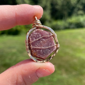 May include: A gold-colored pendant featuring a rough, reddish-purple gemstone. The stone is encased in a wire-wrapped setting, with a loop at the top for a chain. The pendant is held up against a blurred green background.