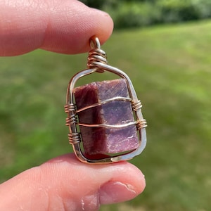 May include: A rectangular, reddish-purple gemstone pendant wrapped in gold-colored wire. The pendant is held up by a gold-colored bail. The background is a blurred green.